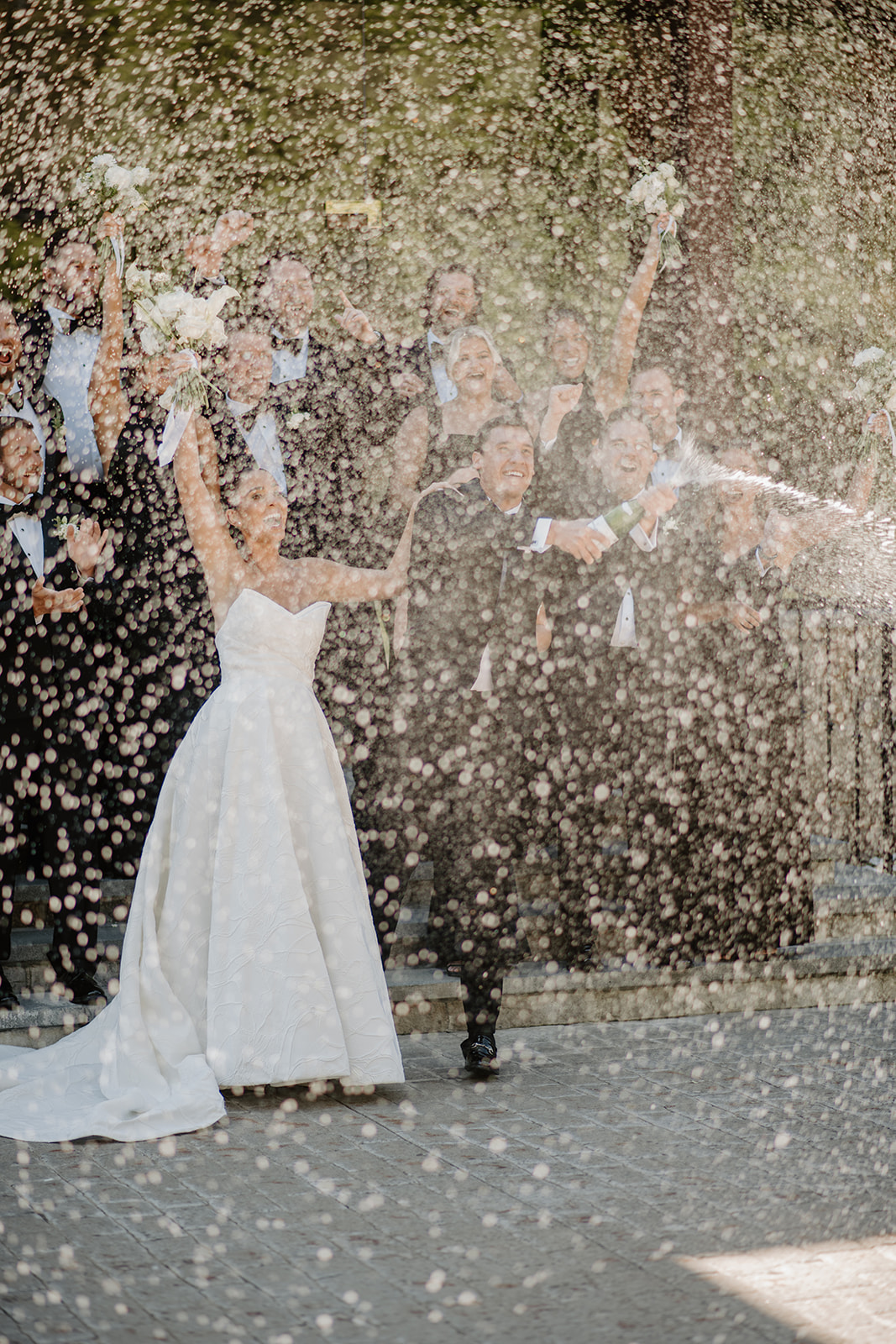 Exuberant bride and groom popping champagne, sprayed in a shower of bubbles with their wedding party cheering.